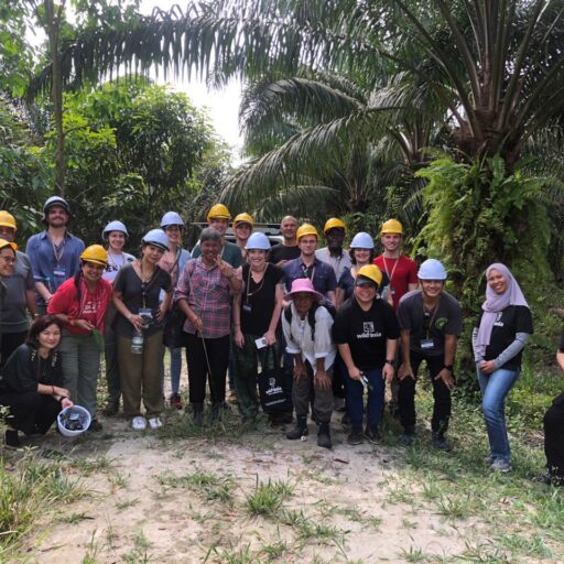 A group photo of participants in the 2022 Wild Asia Smallholder Study Tour in Malaysia, including Conal Judd-English and Judith Murdoch, standing in an oil palm plantation. The group is wearing safety helmets and smiling, surrounded by lush palm trees.